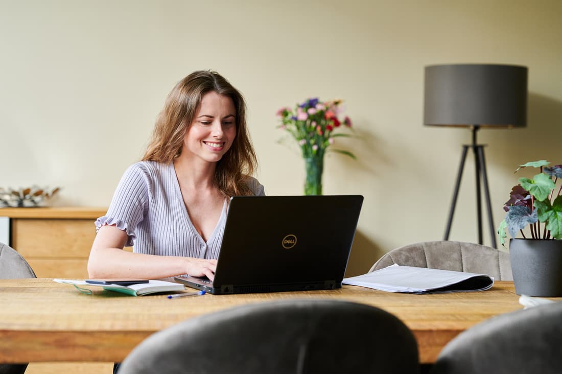 Lachende zorgprofessional zit in haar woonkamer te werken achter haar laptop