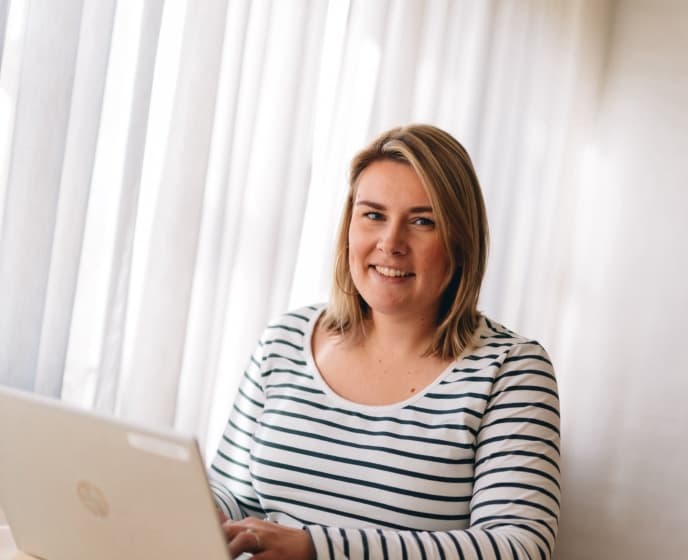 Vrouw met gestreepte trui zit achter een laptop en kijkt glimlachend naar de camera