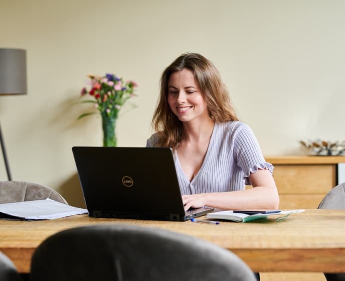 Vrouw werkt glimlachend achter een laptop aan een houten tafel met papieren en een vaas met bloemen